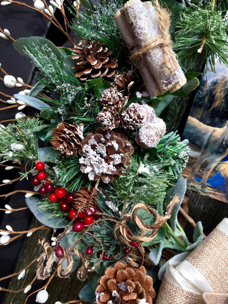 May include: A wintery wreath featuring pine branches, pinecones, red berries, and faux snow. The wreath is decorated with burlap ribbon and a pair of cinnamon sticks tied with twine.