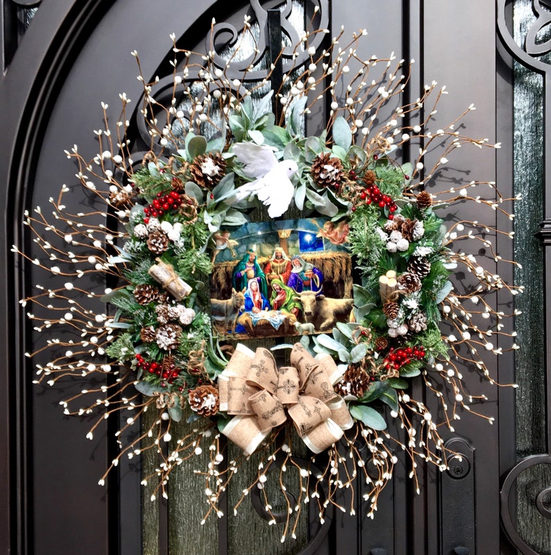 May include: A Christmas wreath featuring a nativity scene at its centre. The wreath is adorned with white berries, pine cones, and greenery. A white dove sits atop the scene.