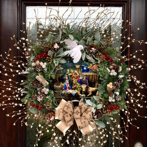 May include: A Christmas wreath featuring a nativity scene in the centre. The wreath is decorated with white berries, pine cones, and a white dove. A brown burlap bow with a cross pattern is tied at the bottom.