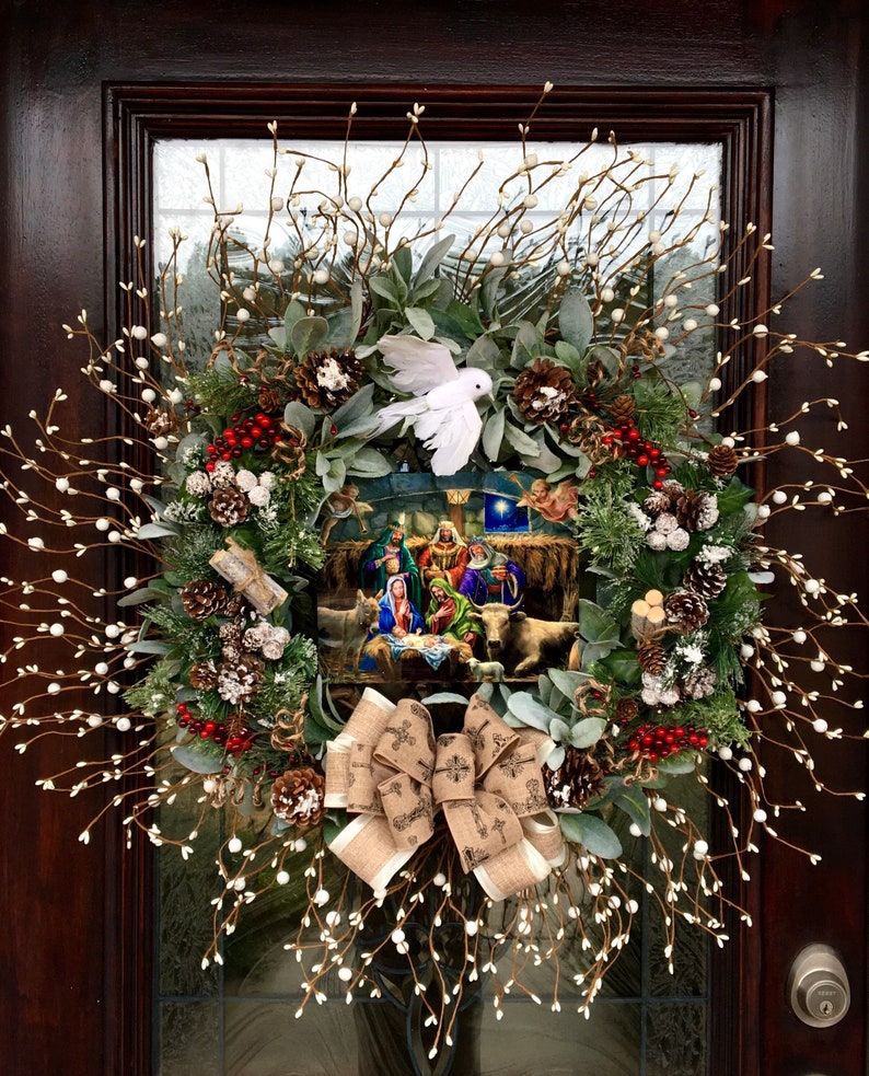 May include: A Christmas wreath featuring a nativity scene in the centre. The wreath is decorated with white berries, pine cones, and a white dove. A brown burlap bow with a cross pattern is tied at the bottom.