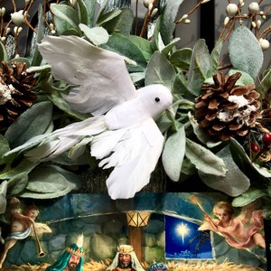 May include: A white dove with its wings outstretched is perched on a wreath of green leaves and white berries. A Christmas card with a nativity scene is visible behind the dove. The card depicts three wise men, a star, and an angel.