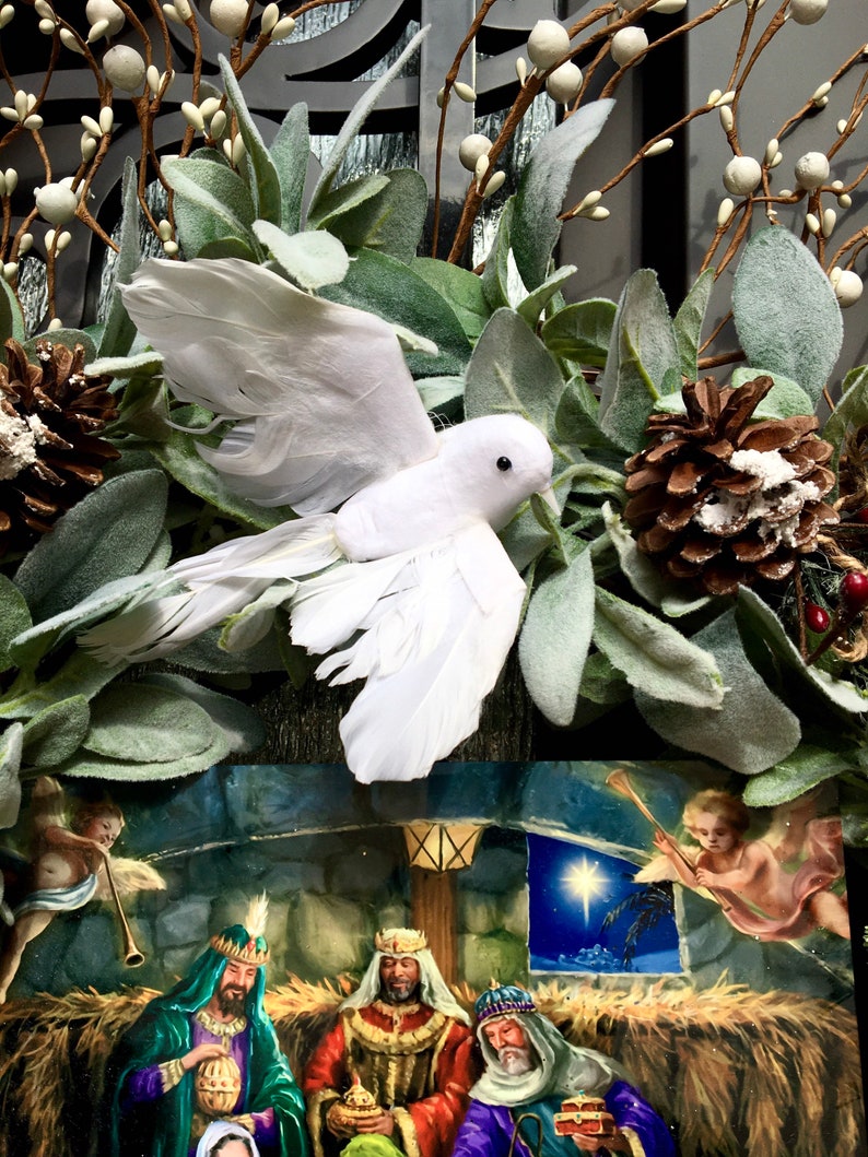 May include: A white dove with its wings outstretched is perched on a wreath of green leaves and white berries. A Christmas card with a nativity scene is visible behind the dove. The card depicts three wise men, a star, and an angel.