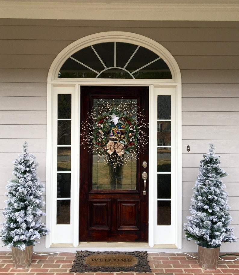 May include: A brown wooden front door with a white wreath decorated with red berries, a white dove, and a gold bow. The door has a glass panel with a decorative arched window above it. Two small, snow-frosted artificial Christmas trees are on either side of the door. A welcome mat is on the ground in front of the door.