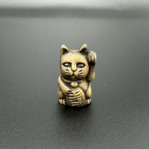 Pode incluir: Uma figurinha de cer&acirc;mica dourada de um gato com a pata direita levantada, uma figurinha tradicional japonesa Maneki-Neko ou "gato da sorte".