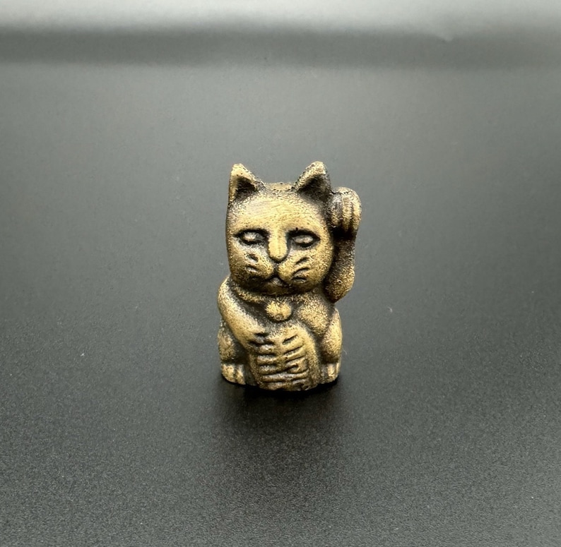 Pode incluir: Uma figurinha de cer&acirc;mica dourada de um gato com a pata direita levantada, uma figurinha tradicional japonesa Maneki-Neko ou "gato da sorte".