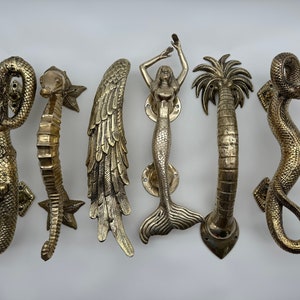 May include: Set of six decorative door handles made of brass. The handles are shaped like a mermaid, a seahorse, a palm tree, a dragon, a wing, and a snake.