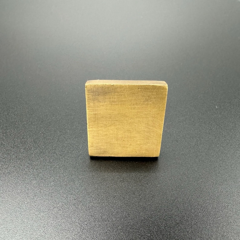 Pode incluir: Uma telha quadrada de lat&atilde;o escovado com uma superf&iacute;cie ligeiramente texturizada.