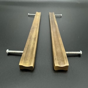 Könnte beinhalten: Zwei Messing-Schubladengriffe mit leicht gekrümmter Form und gebürsteter Oberfläche. Jeder Griff hat an jedem Ende ein einzelnes Schraubenloch.