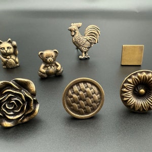 Könnte beinhalten: Sortiment antiker Messing-Schrankknöpfe in verschiedenen Designs, darunter eine Rose, ein gewebtes Muster, eine Sonnenblume, ein Teddybär, ein Hahn und eine Katze.