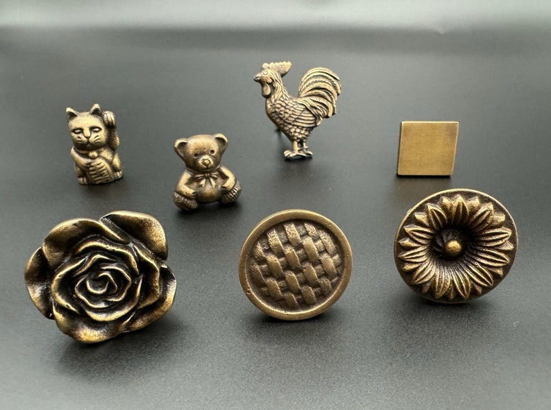 Pode incluir: Conjunto de bot&otilde;es de arm&aacute;rio de lat&atilde;o antigo em v&aacute;rios designs, incluindo uma rosa, um padr&atilde;o tecido, um girassol, um urso de peluche, um galo e um gato.