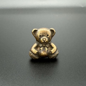 Pode incluir: Uma pequena figura de urso de pel&uacute;cia de cor dourada. O urso est&aacute; sentado com as patas cruzadas e tem um la&ccedil;o de fita em volta do pesco&ccedil;o.