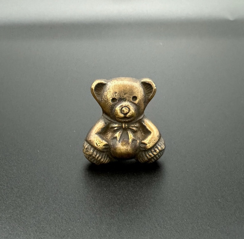 Pode incluir: Uma pequena figura de urso de pel&uacute;cia de cor dourada. O urso est&aacute; sentado com as patas cruzadas e tem um la&ccedil;o de fita em volta do pesco&ccedil;o.