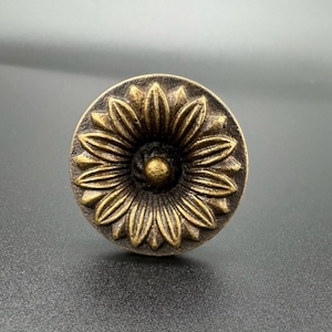 Pode incluir: Uma ma&ccedil;aneta de metal dourado com um design decorativo de girassol. A ma&ccedil;aneta tem um centro elevado e uma superf&iacute;cie texturizada.