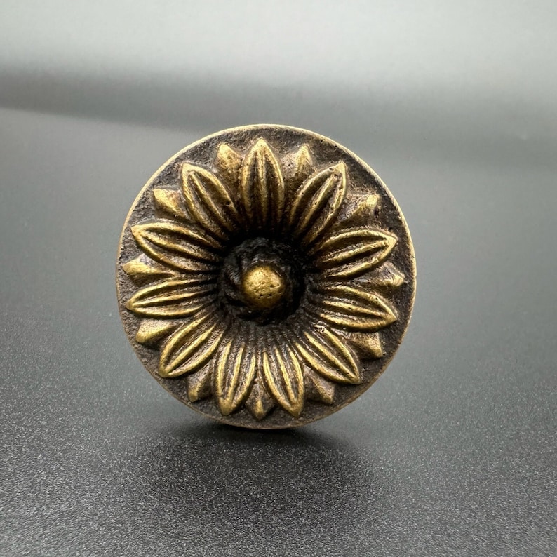 Pode incluir: Uma ma&ccedil;aneta de metal dourado com um design decorativo de girassol. A ma&ccedil;aneta tem um centro elevado e uma superf&iacute;cie texturizada.