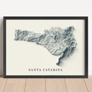 May include: Framed art print featuring a 3D topographic map of Santa Catarina, Brazil. The map is rendered in shades of gray and white, set against a cream-colored background. The title "SANTA CATARINA" is printed in a simple font below the map.