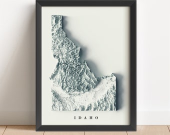 May include: A black framed print of a topographic map of Idaho. The map is in shades of gray and white, with the state's name printed below.