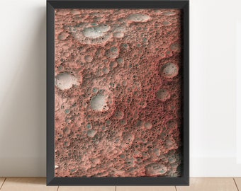 May include: A framed abstract art print with a red, orange, and grey colour scheme. The print features a textured, cratered surface reminiscent of a celestial body.