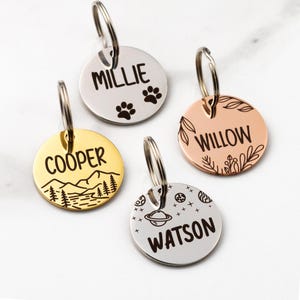 May include: Four personalized pet ID tags with silver, gold, and rose gold finishes. Each tag has a circular shape and includes a name and design. The tags feature the names "Millie", "Cooper", "Willow", and "Watson". Designs include paw prints, mountains, and space-themed graphics.