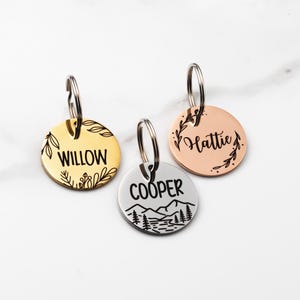 May include: Three round pet ID tags with silver key rings. One is gold with the name "Willow" and floral accents. Another is silver with "Cooper" and a mountain scene. The third is rose gold with "Hattie" and floral accents.
