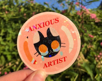 May include: A round sticker with a cartoon cat illustration. The cat is black with white ears and orange eyes, with holographic tears. The sticker has the words "ANXIOUS ARTIST" in red. A pencil illustration is on the left side of the sticker.