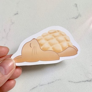 May include: A sticker of a cartoon brown rabbit sleeping on its back with a light brown, round, pastry-like object on its belly. The pastry has a grid pattern on its surface.