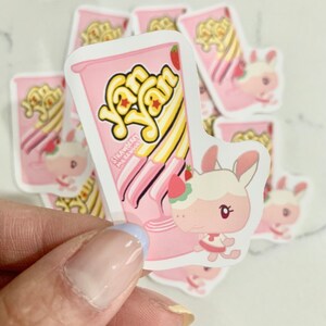 May include: A collection of pink stickers featuring a cartoon bunny and a pink container with the text "Yan Yan". The container has yellow and white stripes and a strawberry on top. The bunny is wearing a pink dress.