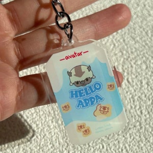 May include: A clear acrylic keychain featuring a cartoon image of Appa, the sky bison from the animated series Avatar: The Last Airbender. The keychain has a blue background with white clouds and the text "Hello Appa" in red letters. The keychain also has the text "_avatar_" in black letters.