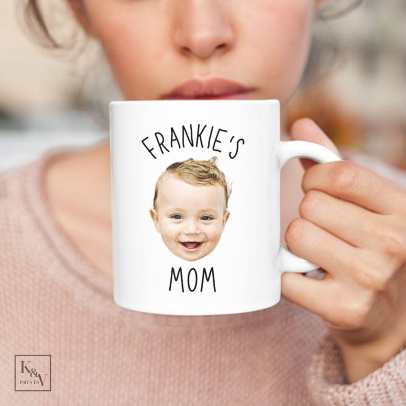 以下が含まれることがあります： 「FRANKIE'S MOM」の文字と笑顔の赤ちゃんの顔がプリントされた白いセラミックマグカップ。 ピンク色のセーターを着た人が持っています。 マグカップには白いハンドルがあり、下部に「K&V PRINTS」のロゴがあります。