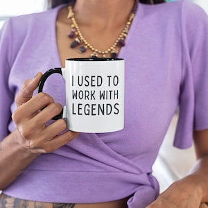 May include: White coffee mug with a black rim and handle. The mug has the text "I used to work with legends" printed in black.