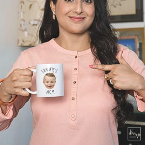 以下が含まれることがあります： 「FRANKIE'S MOM」の文字と赤ちゃんの顔がプリントされた白いセラミックマグカップ。ピンクのシャツを着た人が持っています。パーソナライズされたギフトです。