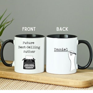 May include: White ceramic mug with a black rim and handle. The front of the mug features a black and white illustration of a typewriter and the text "Future Best-Selling Author". The back of the mug has the text "Daniel" written in black with a hand holding a pen above it.