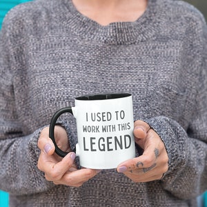 Op de afbeelding: Een witte koffiemok met een zwarte handgreep en een zwarte rand. De mok heeft de tekst "I used to work with this LEGEND" in zwart gedrukt.