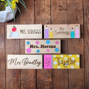 May include: Wooden teacher name plaques with various designs. One features an apple and books, another pencils, and a third with flowers. Other plaques include the names Mrs. Scruggs, Mrs. Cummings, and Mrs. Moreno.