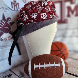 May include: A red and white patterned scrub hat with a black tie, featuring the Texas A&M logo and other team graphics. A football and basketball are in the background.