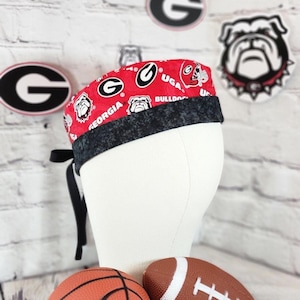 May include: A red and black patterned scrub cap featuring the University of Georgia Bulldogs logo and text. The cap has a black band and ties. The background includes Georgia Bulldogs logos and a football and basketball.