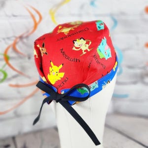 May include: A red surgical scrub cap featuring various cartoon characters and text. The cap has a blue trim and a black bow. The characters include Pikachu, Meowth, and Bulbasaur. The cap is designed to be worn during medical procedures.