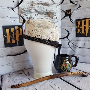 May include: A brown and black fabric hat with a Harry Potter themed design. The hat has a black ribbon with gold accents and the words "Time &  Space" and "Marauder's Map" printed on it. The hat is sitting on a white mannequin head. There is a black mug with the Harry Potter crest on it and a brown wooden wand in the image.