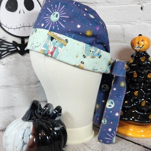 May include: A blue and white patterned surgical scrub hat with a tie. The hat features a skull and crossbones design and the words "Happy" and "Boo!" The tie is also blue and white with a skull and crossbones design.