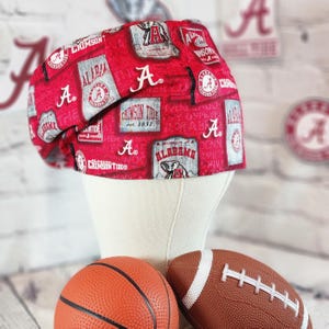 May include: A red and white patterned scrub cap featuring the University of Alabama logo and the words "Crimson Tide". The cap is displayed on a mannequin head with a basketball and a football in the foreground.