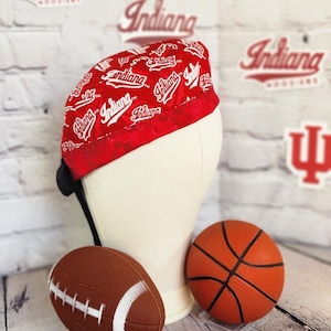 May include: Red and white Indiana-themed head covering with the word "Indiana" repeated across the fabric. A black strap is attached to the head covering. A brown football and orange basketball are in the foreground.