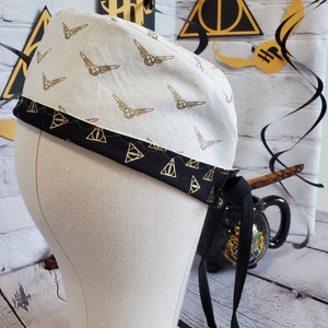 May include: A white fabric scrub hat with a black trim and a gold pattern of snitches and golden snitch wings. The hat has a black tie that is tied around a black cauldron.