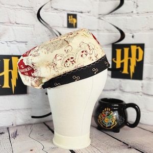 May include: A cream-colored, patterned hat with a black brim featuring gold-colored glasses. The hat displays a map design with red accents and the text "The Marauder's Map". A black mug with a gold crest is also visible.