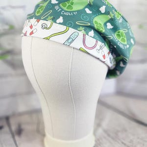 May include: A scrub cap featuring a dark green upper section with a repeating pattern of green gallbladder illustrations, white clouds, and the text HOLY CHOLEY! The white band displays a medical print with colorful pills, stethoscopes, and bandages.