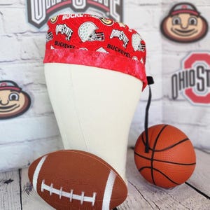 May include: A red scrub cap with a white Ohio State Buckeyes logo and football helmets. A brown football and orange basketball are in the foreground. The cap is on a mannequin head.