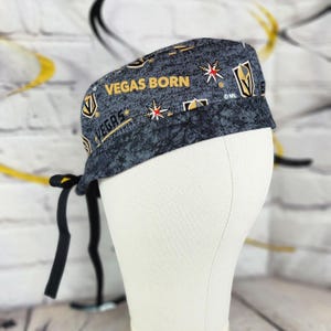 May include: A gray and black patterned cap with the words "VEGAS BORN" and "VEGAS" in gold lettering. The cap features gold and red starburst designs and a black tie closure. The cap is on a white mannequin head.