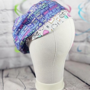 May include: A scrub cap featuring a purple background with scattered medical text and a white band decorated with various medical illustrations including stethoscopes, hearts, and pill bottles. The fabric displays phrases like STAT, NPO TIME, and SUTURE.