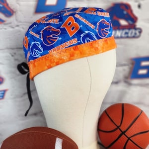 May include: A blue and orange Boise State Broncos scrub cap. The cap features the team's logo and the words "Broncos" and "Boise State" in white and orange. The cap has an orange brim and black ties.