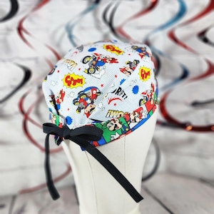 May include: A light blue scrub cap featuring a colorful print of cartoon characters in action. The cap has a black bow and ties. The design includes comic book style speech bubbles and the word "DRIFT!"