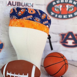 May include: A blue and orange Auburn Tigers scrub cap with a repeating logo pattern. The cap is displayed on a mannequin head, with a football and basketball in the foreground. The background includes Auburn Tigers logos.