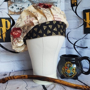 May include: A white and brown fabric scrub hat with a black and gold patterned band. The hat features a map design with the words "Marauder's Map" printed on it. The hat is sitting on a white mannequin head. A black mug with a Hogwarts crest is in the background.